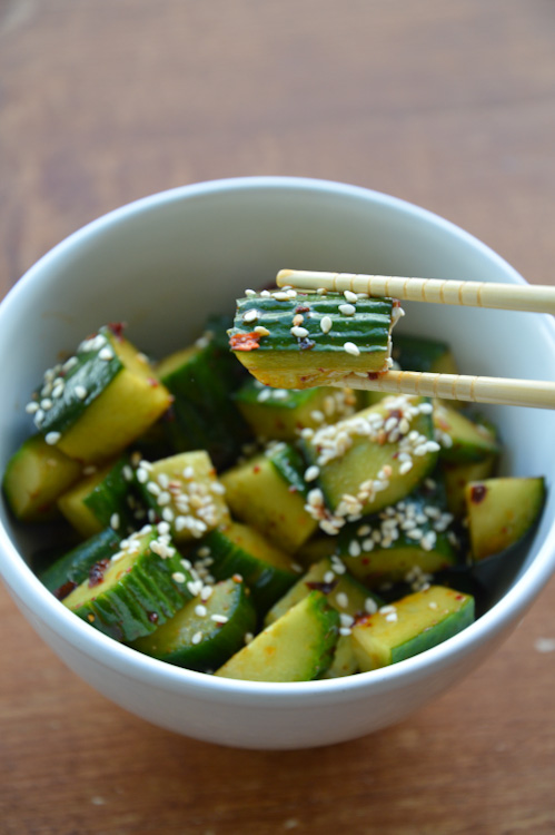 White bowl of Sichuan cucumber salad topped with sesame seeds; chopsticks holding one piece of cucumber