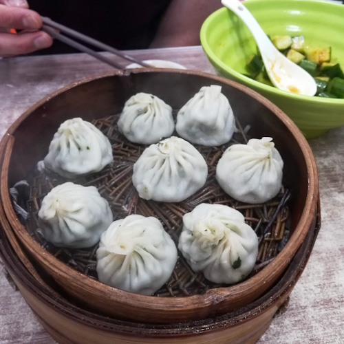 A basket of steamed soup dumplings and a green bowl of cucumber salad in Shanghai