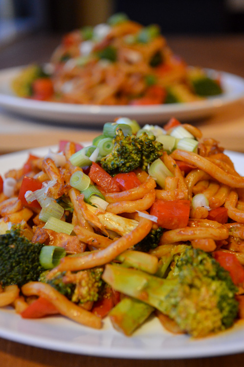 Two plates of veggie udon noodles with peanut sauce featuring broccoli, red pepper, and tofu