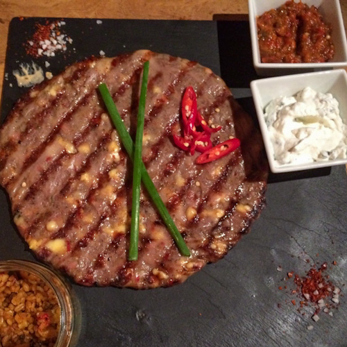 Pljeskavica (Serbian hamburger) on a black plate surrounded by toppings, eaten in Warsaw