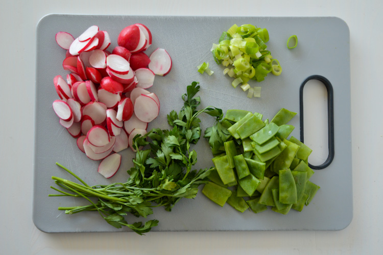 Grey cutting board with: sliced radishes, a bunch of parsley, chopped snow peas, and minced scallions