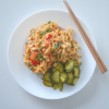White plate of spicy peanut noodles with cucumber salad and chopsticks on a white background