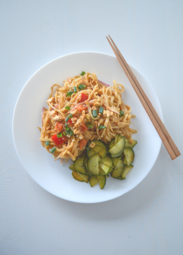 White plate of spicy peanut noodles with cucumber salad and chopsticks on a white background