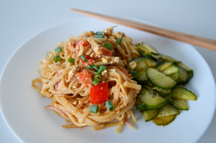 Angled view of a white plate of spicy peanut noodles with red peppers next to pickled cucumbers, with chopsticks