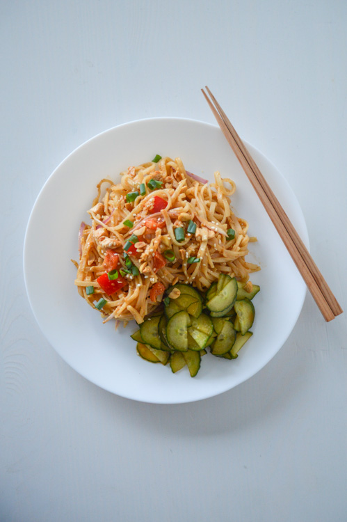 White plate with spicy peanut noodles, served cold, and a side of pickled cucumbers, seen with chopsticks