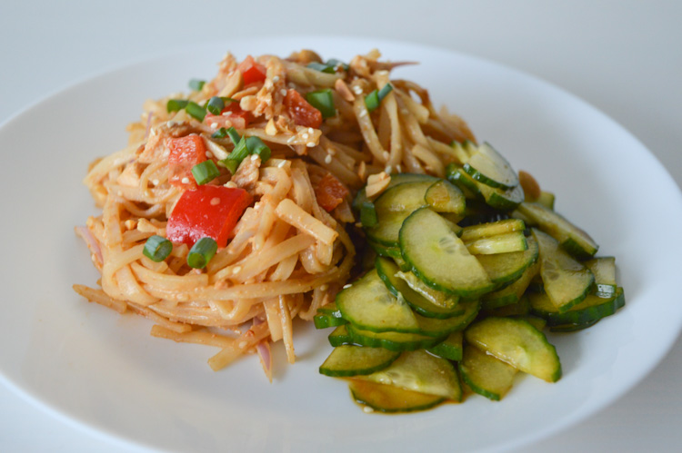 A white plate of cold noodles with red peppers, chicken, topped with a spicy peanut sauce served with cucumbers on the side