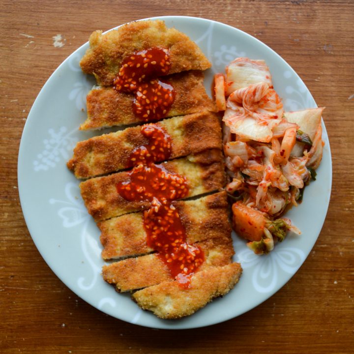 Sliced piece of fried, thin meat with gochujang sauce on top served with a side of kimchi