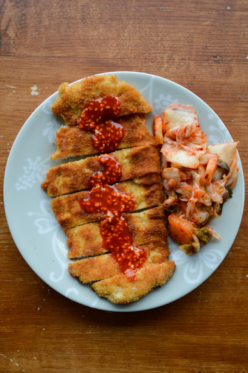 Fried pork cutlet sliced, covered in red gochujang sauce, with a side of kimchi on a plate