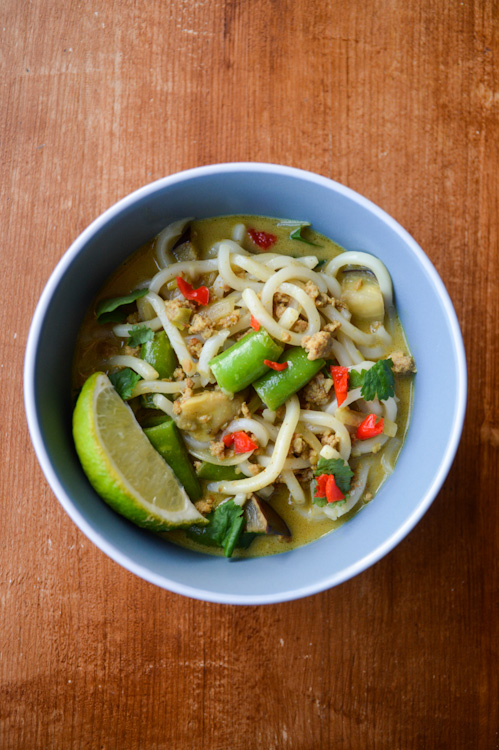 Blue bowl of khao soi curry udon with sugar snap peas, spicy red peppers, green herbs, and a lime wedge