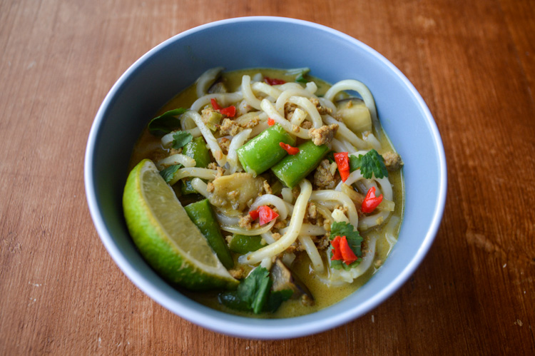 Blue bowl of khao soi curry udon with sugar snap peas, spicy red peppers and a lime wedge