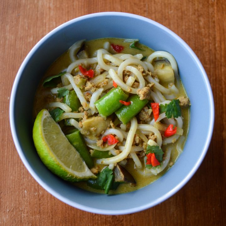 Close up of a blue bowl of curry udon with sugar snap peas, spicy red peppers and a lime wedge