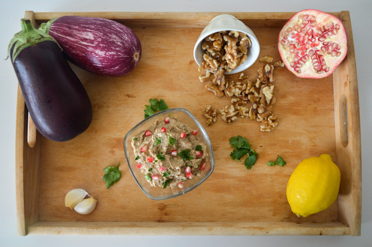 A wood tray with a dish of pkhali and all its ingredients: eggplant, garlic, walnuts, cilantro, lemon, and pomegranate