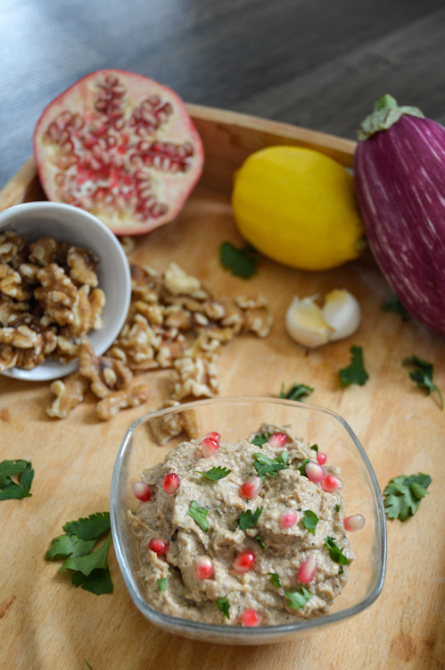 A wooden tray with a dish of eggplant pkhali, walnuts, half a pomegranate, a lemon, an eggplant, and garlic cloves