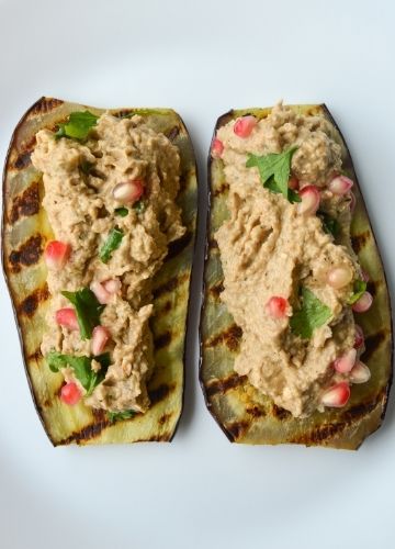 Two pieces of grilled eggplant topped with eggplant pkhali with pomegranate seeds and green herbs on a white plate