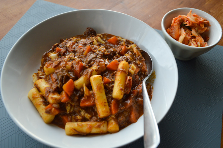 Close up of a bowl of Korean beef stew with a spoon, and a small side bowl of kimchi