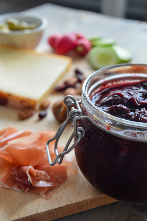 Close up of glass jar of plum chutney sitting on a wood board with other borrel snacks (meat, cheese, nuts, etc.)