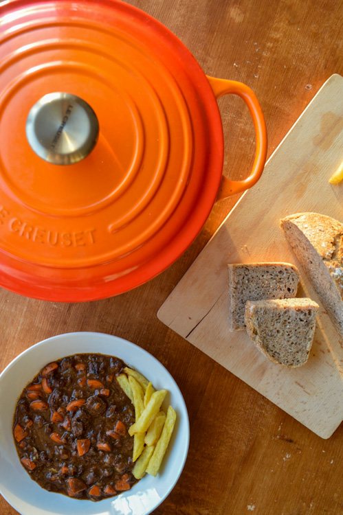 Overhead shot of a bowl of beef stew with fries, an orange Dutch oven, and a wood board with bread