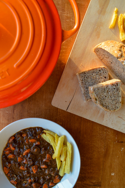 Overhead shot of a bowl of stoofvlees Flemish beef stew, an orange Dutch oven, and a wood board with bread and fries