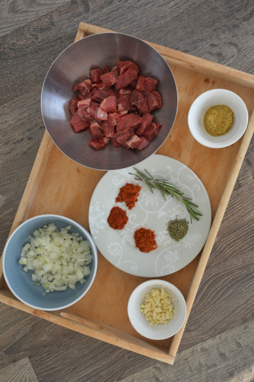 Overhead shot of stoofvlees ingredients including cubed beef, minced onions, minced garlic, mustard, herbs, and spices