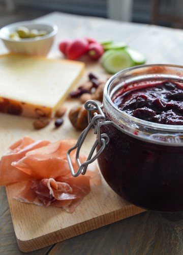 Glass jar of spiced plum chutney half out of frame along with a board of meats and cheeses