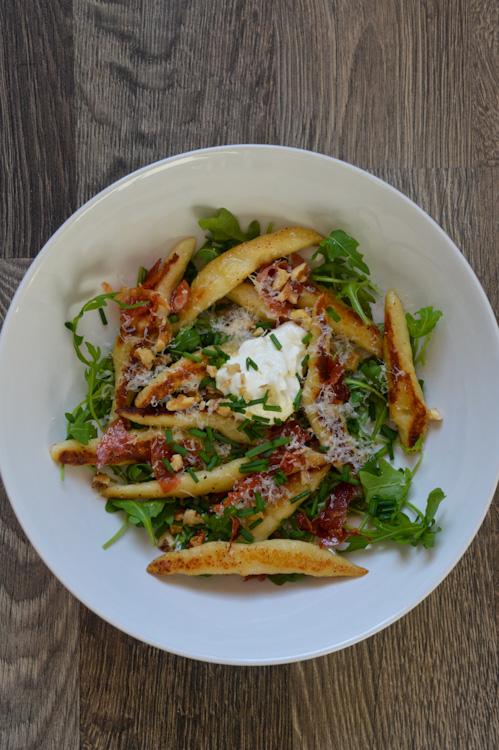 A white bowl with Schupfnudeln(handmade potato dumplings), arugula, crispy ham, chopped walnuts, and sour cream