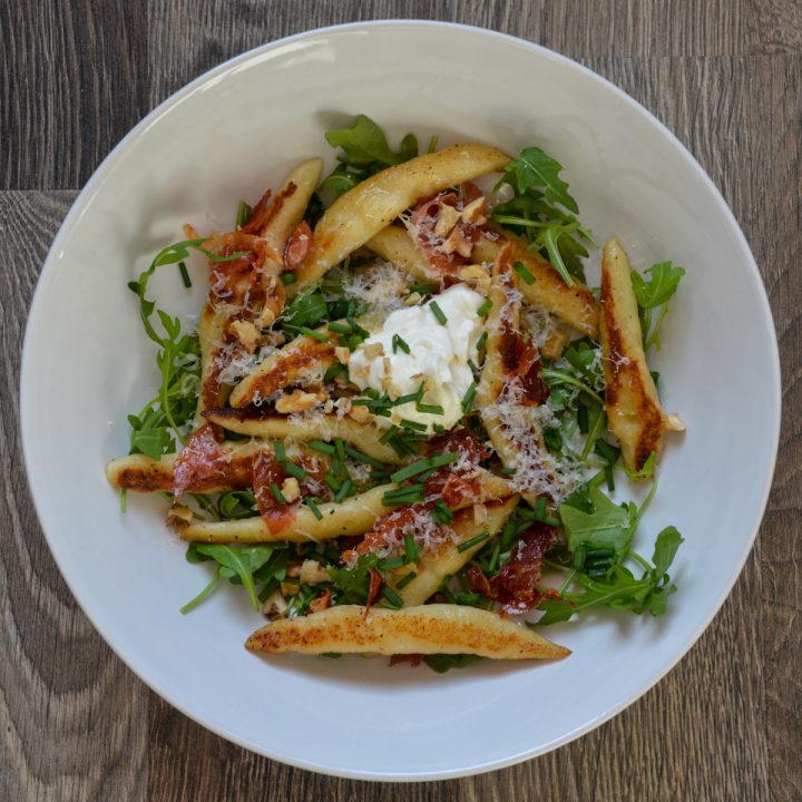 White bowl with cylindrical handmade dumplings, arugula, crispy ham, chopped walnuts, and sour cream