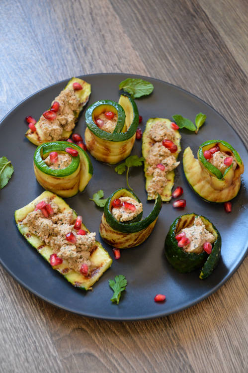 Black plate with 3 pieces of zucchini topped with Georgian walnut paste and 5 zucchini spirals with the paste in the center, all decorated with fresh green herbs and pomegranate seeds