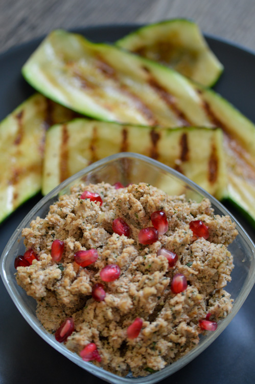 Georgian walnut spread topped with pomegranate seeds in a glass bowl in the foreground; behind it are a few pieces of grilled zucchini