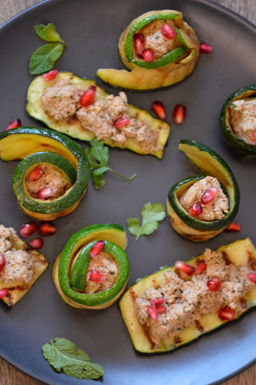 Black plate with zucchini topped with and wrapped around Georgian walnut paste in spirals; plate is artfully decorated with fresh herbs and pomegranate seeds