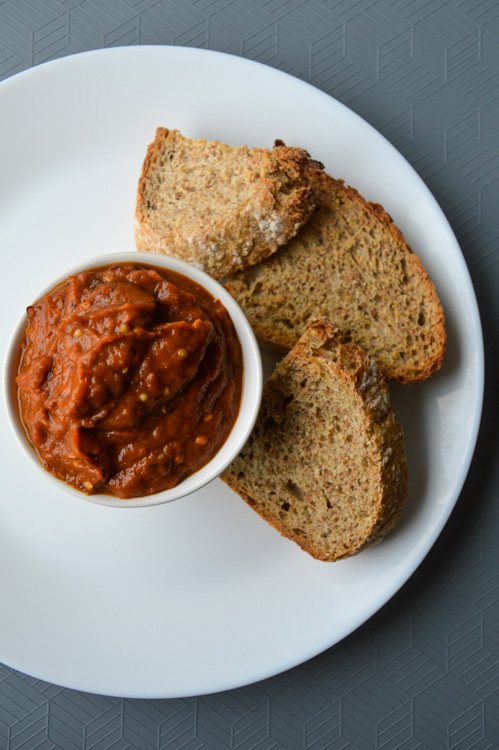 A white plate with a small bowl of zacusca and three pieces of sliced wheat bread