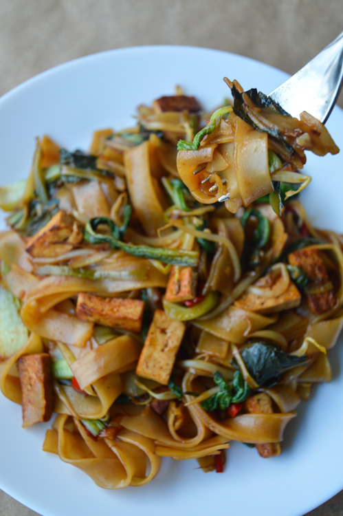 White plate full of brown wide rice noodles, tofu, and veg; a fork hovers over holding a tasty looking bite