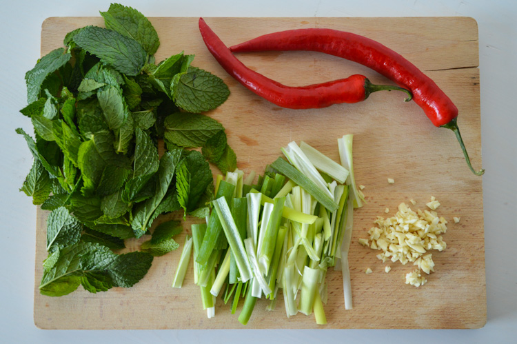 Wood board with a pile of mint leaves, julienned scallions, long red peppers, and minced garlic