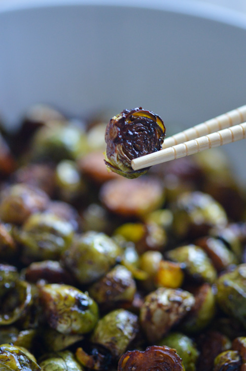 A single, browned Brussels sprout held above a bowl of its brethren with light colored chopsticks