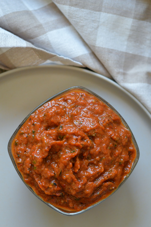 Square glass bowl of roasted vegetable ajvar (a beautiful red pepper and zucchini spread) on a beige plate on a beige and white checked tea towel