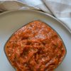 Square bowl of roasted vegetable ajvar (a beautiful red pepper and zucchini spread) on a beige plate on a beige and white checked tea towel