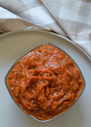 Square bowl of roasted vegetable ajvar (a beautiful red pepper and zucchini spread) on a beige plate on a beige and white checked tea towel