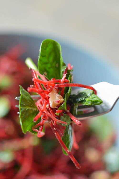 In the foreground, a tasty bite of beet spinach salad on a fork; in the background, a slightly blurry bowl of the rest of the salad in the background