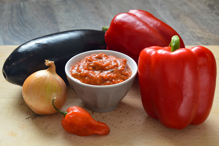 A small bowl of zacusca on a wooden board surrounded by 2 red bell peppers, 1 small spicy red pepper, 1 onion, and 1 eggplant